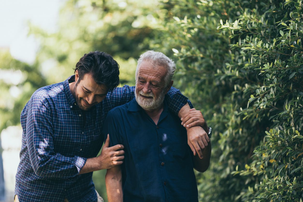 The old man and his son are walking in the park. A man hugs his elderly father. They are happy and smiling